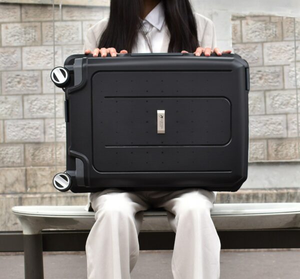 Valise rigide Airtex noire, tenue à la main, posée sur un banc en pierre